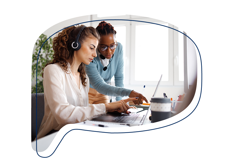 Zwei Frauen mit Headsets arbeiten gemeinsam an einem Laptop – eine sitzt am Schreibtisch, die andere steht daneben und zeigt auf den Bildschirm.