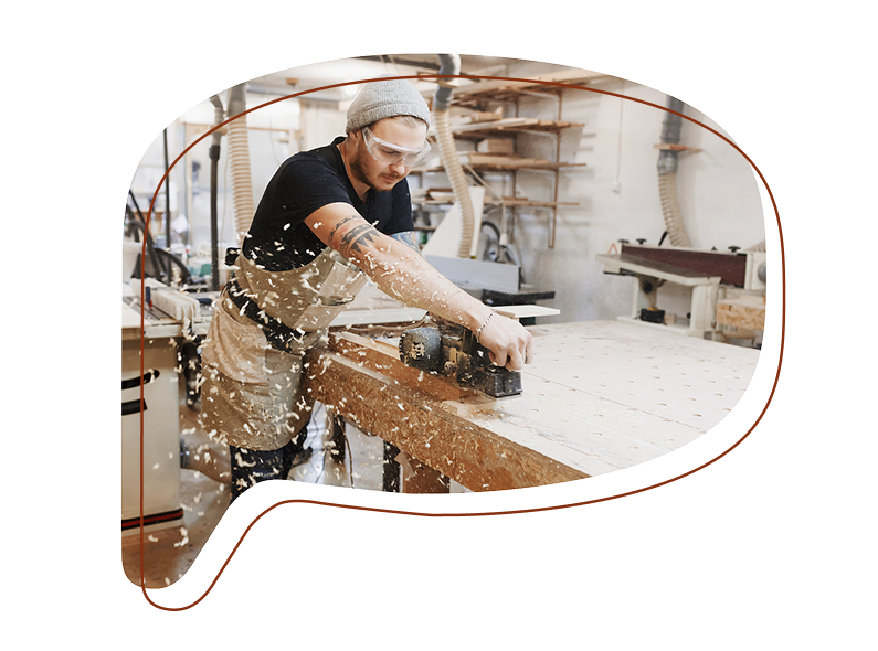 Ein Mann mit Schutzbrille und Schürze hobelt in einer Schreinerei ein Holzbrett – Späne fliegen durch die Luft.